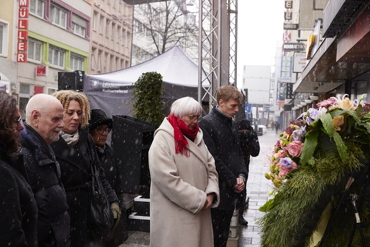 Winterlich gekleidete Frauen und Männer stehen im Gedenken vor einem mit Blumen geschmückten Trauerkranz