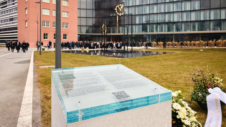 Gläserne Gedenktafel mit Betonsockel am ErinnerungsortMünchen-Riem 1970. Im Hintergrund das Kunstwerk von Alicja Kwade und der ehemalige Flughafentower, die Brainlab Firmenzentrale.