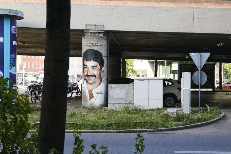 Fotografie eines Graffiti-Porträt von Theodoros Boulgarides, das auf einen Brückenpfeiler gesprüht wurde. Unter der Brücke sind geparkte Autos zu erkennen, davor verläuft eine Straße.