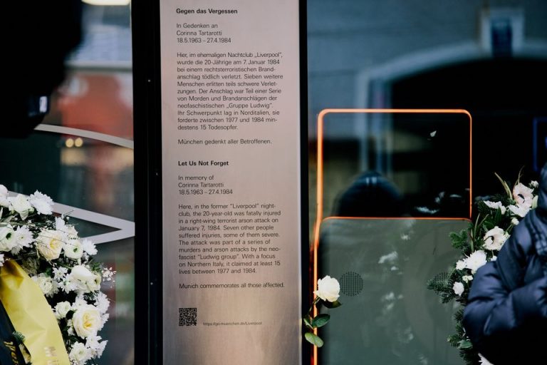 Bild einer Gedenkstele aus Edelstahl mit schwarzer Inschrift, die vor einer gläsernen Ladenfront aufgestellt ist. An beiden Seiten der Stele sind Teile von Gedenkkränzen zu erkennen.