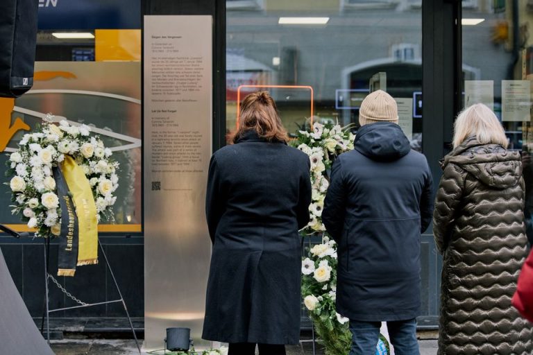 Eine Gedenkstele aus Edelstahl mit schwarzer Inschrift ist auf dem Gehsteig vor einer Ladenfront aufgestellt. Neben der Stele sind drei Gedenkkränze mit weißen und gelben Blumen platziert. Drei Personen stehen vor der Stele, ihre Rücken sind der Fotografierichtung zugewandt.