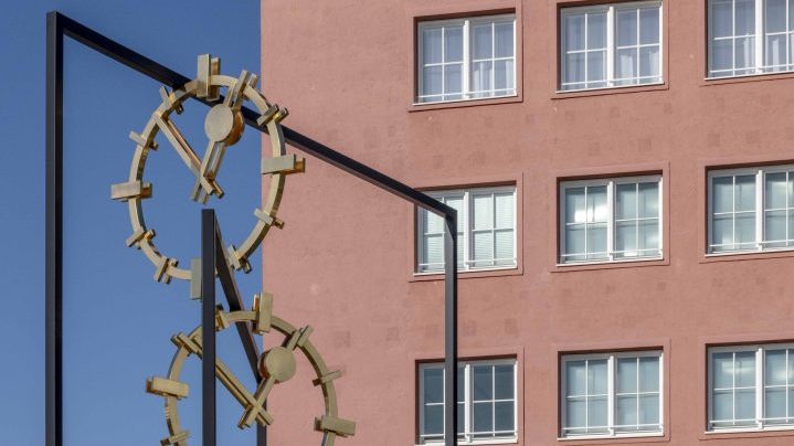 Stahl und Bronzeskulptur aus Rahmen und Zifferblättern, im Hintergrund eine Gebäudefassade aus den 1930er Jahren.
