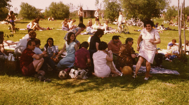 Mobile Slider image "City is Migration". Colour photograph of a group of people sitting on picnic blankets in a park. A woman from the group, wearing a pink summer dress, has stood up and is laughing at the camera.