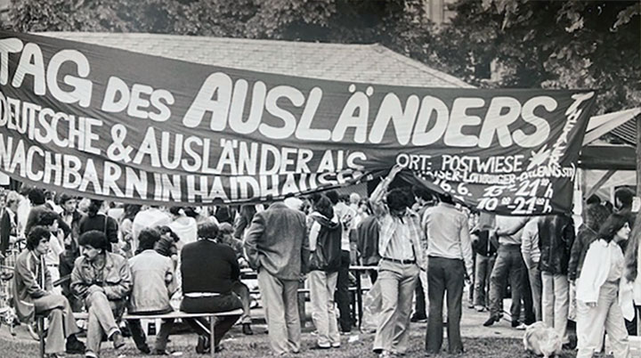 Menschen stehen und sitzen in einem Park und blicken in Richtung eines Transparents mit der Aufschrift: "Tag des Auslängers"