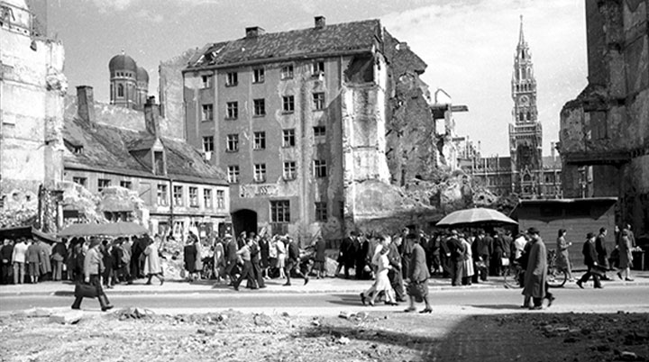 Schwarzweißfoto einer historischen Stadtansicht, leicht bewölkter Himmel. Die Gebäude sind stark beschädigt, teils sind es Ruinen oder Überreste abgerissener Gebäudeteile. Links hinter einer zerstörten Gebäudefassade ragen die Türme der Münchner Frauenkirche hoch. In der rechten Bildhälfte ist ein Gebäude vollkommen zerstört. In dieser Lücke ragt zwischen halbzerstörten Grundmauern der Turm des Neuen Münchner Rathauses empor. Im Vordergrund Geröll. Mehrere Grüppchen von überwiegend erwachsenen Menschen sind in Bewegung oder drängen sich um improvisierte Verkaufsstände unter Planen und Schirmen. Die meisten tragen lange Kleidung oder lange Strümpfe. Die Fotografie zeigt das Rosental in München nach Ende des zweiten Weltkriegs.