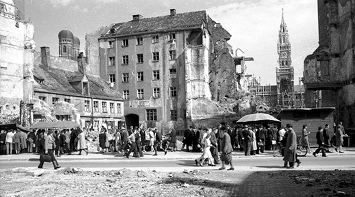 Schwarzweißfoto einer historischen Stadtansicht, leicht bewölkter Himmel. Die Gebäude sind stark beschädigt, teils sind es Ruinen oder Überreste abgerissener Gebäudeteile. Links hinter einer zerstörten Gebäudefassade ragen die Türme der Münchner Frauenkirche hoch. In der rechten Bildhälfte ist ein Gebäude vollkommen zerstört. In dieser Lücke ragt zwischen halbzerstörten Grundmauern der Turm des Neuen Münchner Rathauses empor. Im Vordergrund Geröll. Mehrere Grüppchen von überwiegend erwachsenen Menschen sind in Bewegung oder drängen sich um improvisierte Verkaufsstände unter Planen und Schirmen. Die meisten tragen lange Kleidung oder lange Strümpfe. Die Fotografie zeigt das Rosental in München nach Ende des zweiten Weltkriegs.